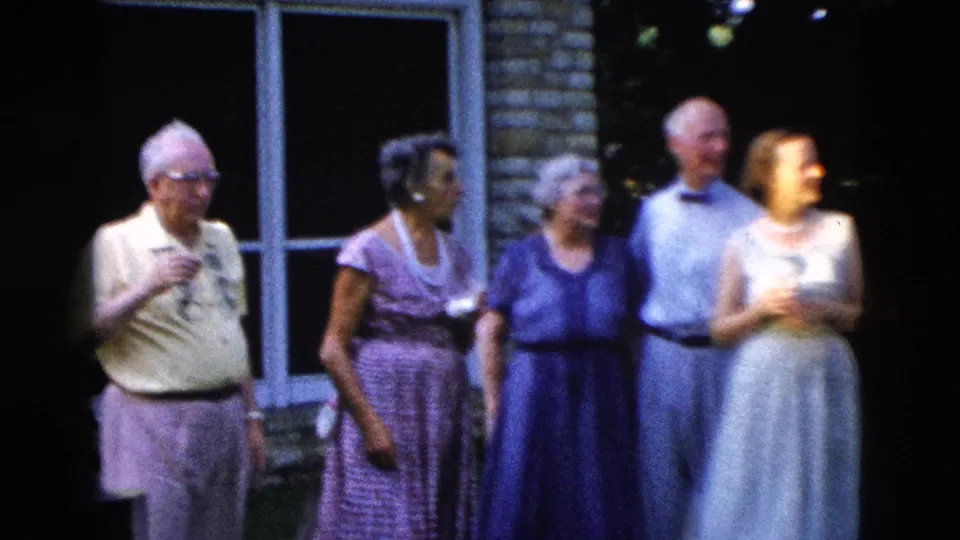 Four older adults — a man with glasses holding a drink, women in coordinating blue and purple dresses — converse at a backyard gathering, circa 1958.