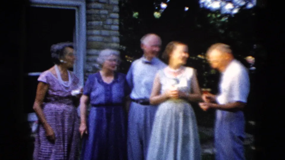 Five older adults — two men in light shirts, women in coordinating purple and blue dresses — stand together conversing in a backyard, circa 1958 summer gathering.