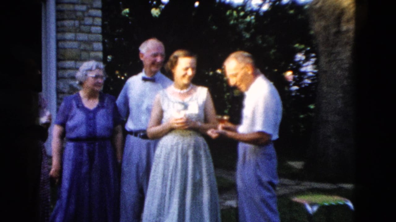 Four adults in 1950s clothing stand outside a stone house, gathered in conversation in a garden.