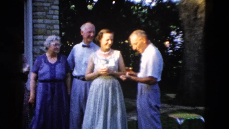 Group of four adults in late-1950s dress — two women in blue and grey, two men in dress shirts — conversing in a backyard garden beside a stone house, circa 1958.