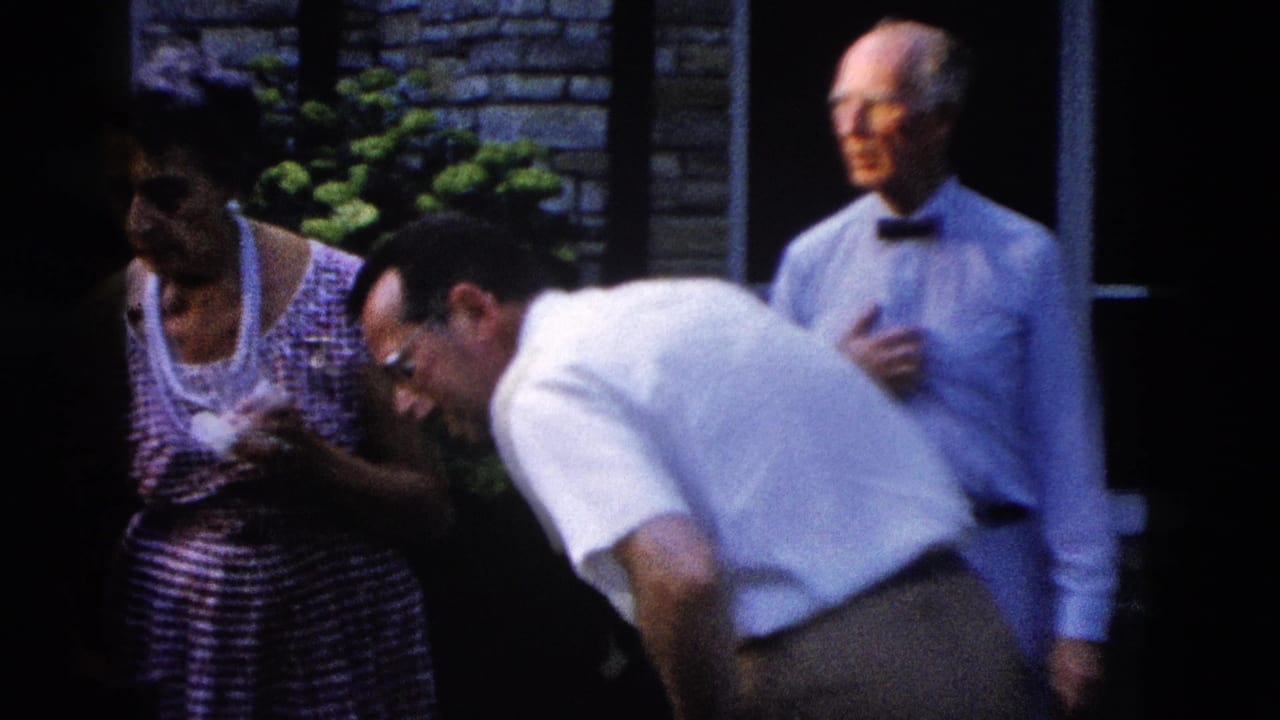 Man in white shirt leans forward laughing beside a woman in a striped dress and an older man in a bow tie.