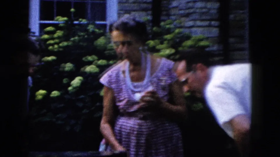 Close shot of a woman in a mid-century striped dress examining a small object with a man leaning in, lush garden shrubs visible in the background, circa 1958 Wyoming.