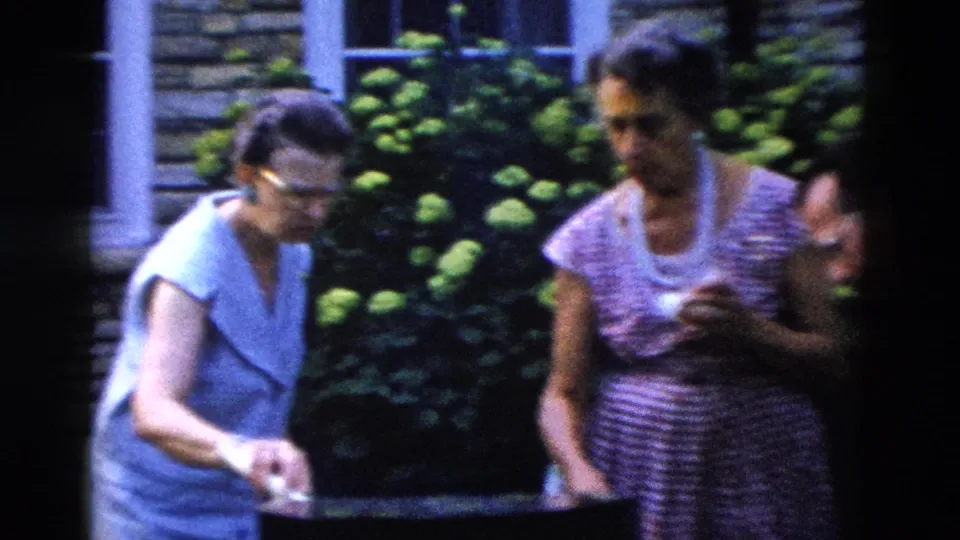 Two adult women in late-1950s summer clothing stand together in a backyard garden, both looking downward, stone wall and dense flowering shrubs visible behind them.