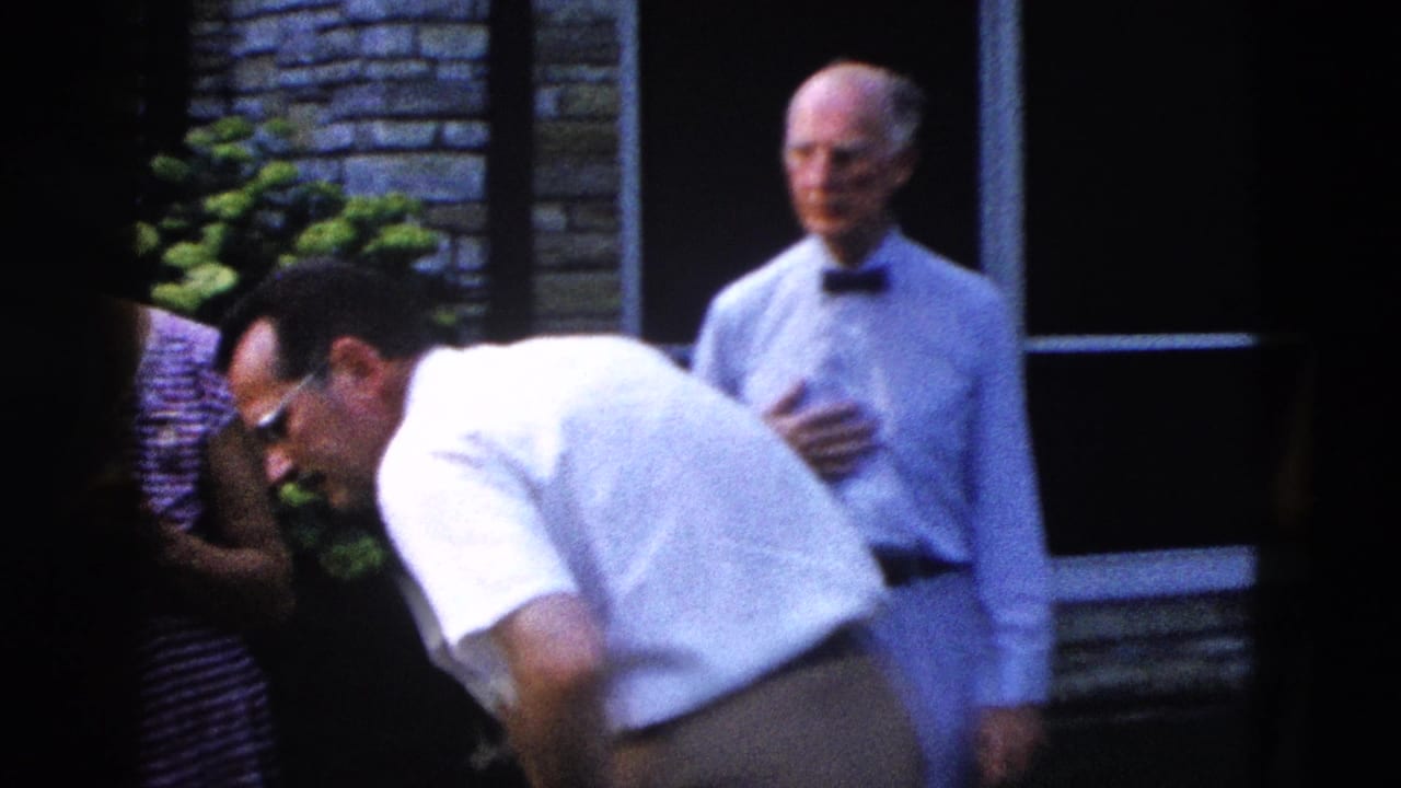 Two men outdoors — one in white shirt bending forward, one older man in bow tie standing — against a stone brick wall.