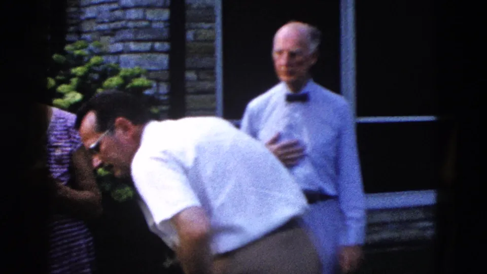 Two men at an outdoor gathering, circa 1958; younger man leans forward while an older man in bow tie and light blue shirt stands near a stone brick building.