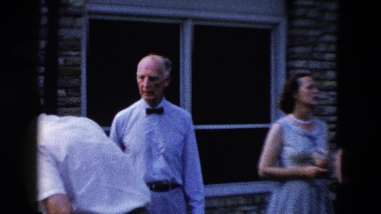 Older man in light blue shirt and bow tie stands outdoors; woman in sleeveless dress at right holds a drink, stone wall and screened window behind them.