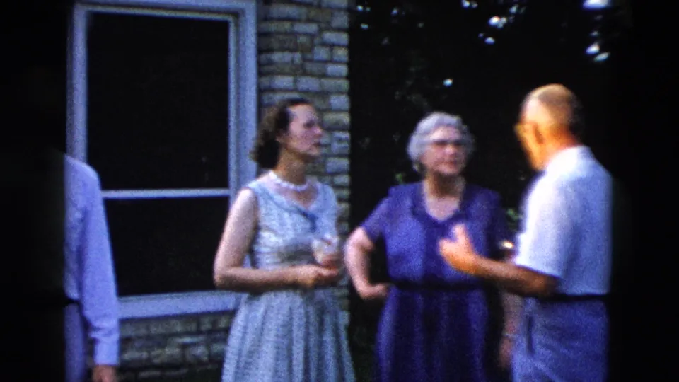 Three adults at an informal outdoor gathering, circa 1958; two women in mid-century shift dresses, one with a pearl necklace, converse with a man in a white shirt.