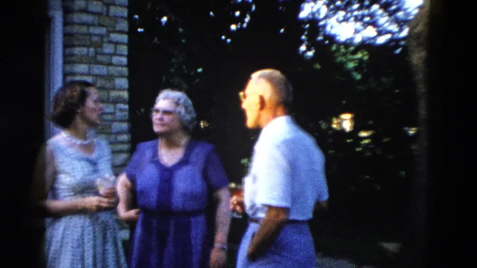 Three adults at an outdoor social gathering circa 1958; women in period dresses, man in white short-sleeve shirt, stone building facade and hydrangeas behind.