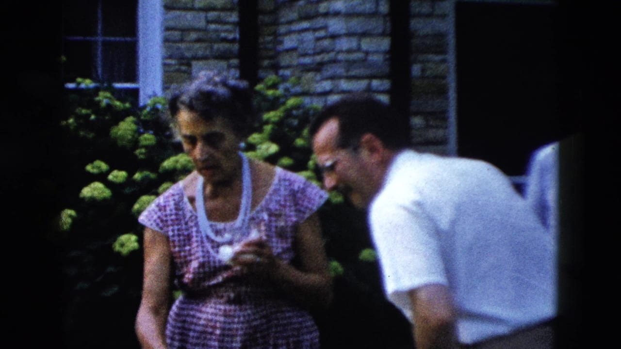 Close shot of two adults at a 1950s outdoor gathering circa 1958; woman in pink patterned dress holds a small cup, man in white shirt wears horn-rimmed glasses.
