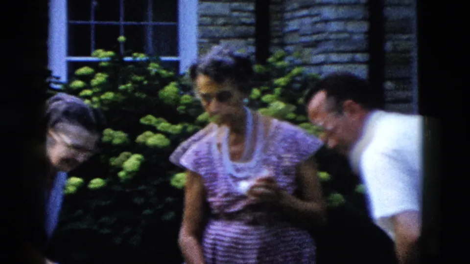 Three adults at a 1950s backyard gathering circa 1958; woman in pink checked dress holds cup, woman in blue partially visible at left, man in white shirt at right, hydrangeas behind.