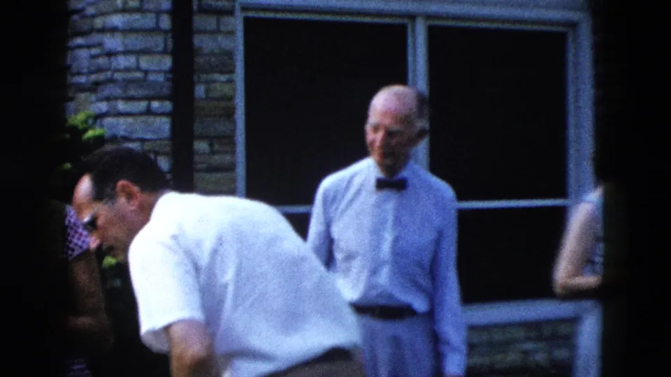 Two older men at a late-1950s outdoor gathering — one with dark-rimmed glasses bending forward, the other in a white shirt and bow tie — standing near a brick house with multi-pane windows.