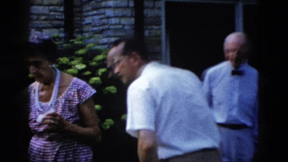 Woman in pink gingham sleeveless dress and pearl necklace in foreground; man in white shirt leaning forward mid-ground and older man behind, all near large flowering shrubs at a 1950s backyard gathering.