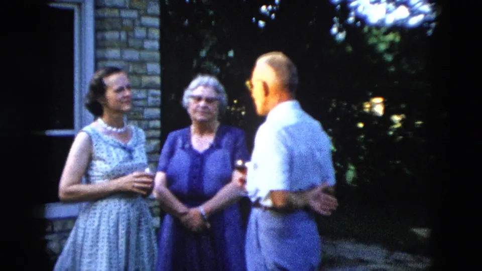 Three adults in backyard conversation beside a stone-clad house, from what appears to be a 1958 American home movie shot on 8mm film.