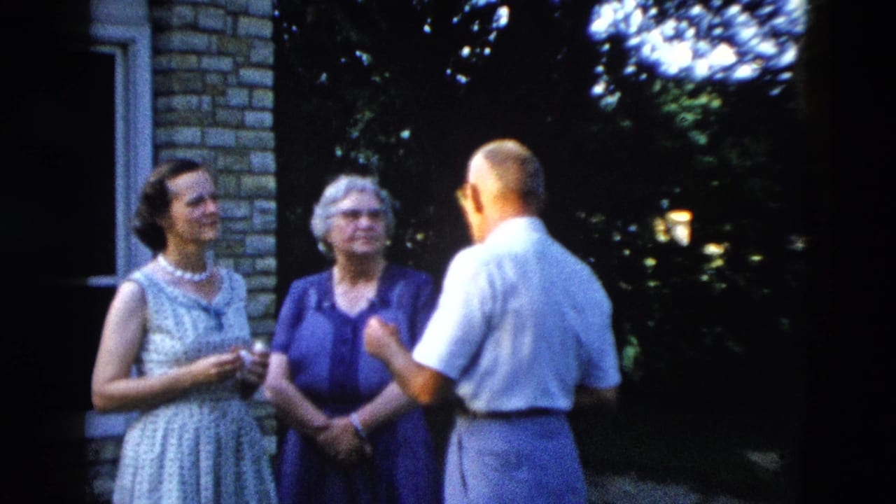 Three adults in animated outdoor conversation beside a stone-clad house, with the man gesturing, from a 1958 American 8mm home movie.
