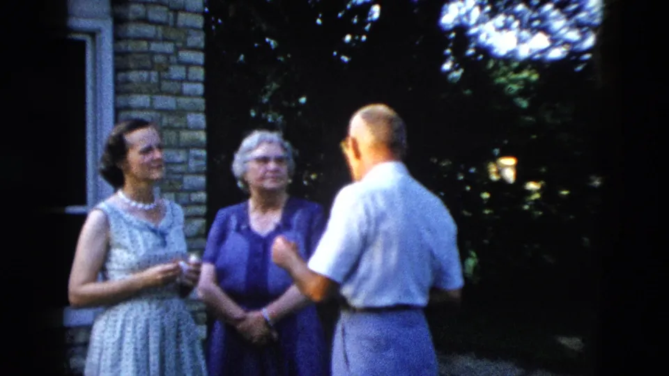 Three adults in animated outdoor conversation beside a stone-clad house, with the man gesturing, from a 1958 American 8mm home movie.