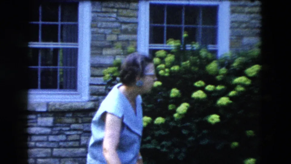 A woman in a light blue top stands in profile before a stone house wall and a full hydrangea shrub in bloom, from a 1958 American backyard home movie.