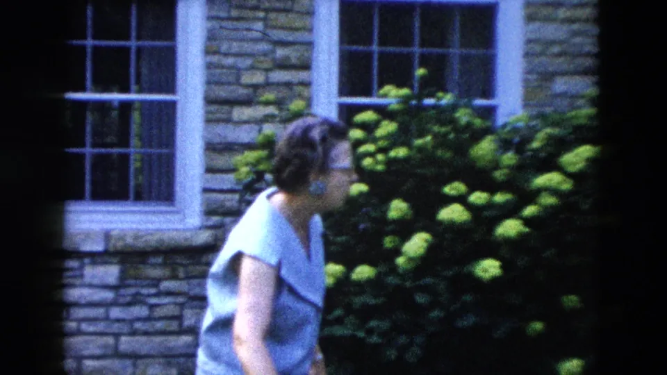 A woman in a light blue top turns slightly, framed against a stone house wall with white double-hung windows and a full hydrangea shrub, from a 1958 home movie.