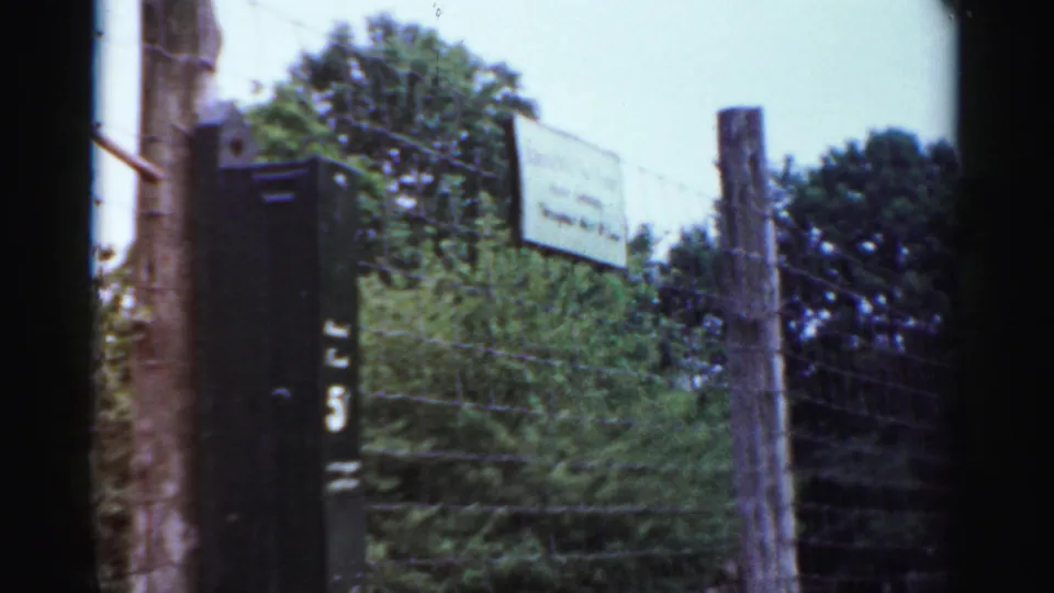 The camera frames a wire fence, wooden post, and small sign at the Catskill Game Farm in 1957, with foliage behind the enclosure.