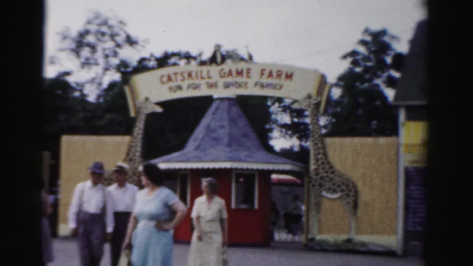 1957 Catskill, New York visitors approach the Catskill Game Farm entrance, with an arched sign, giraffe figures, and a red ticket booth visible.