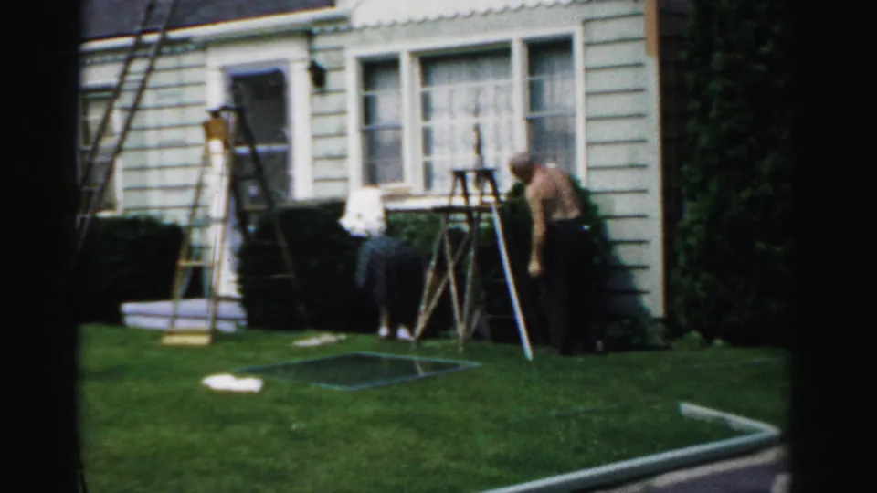 A backyard home-repair scene shows a shirtless man and a woman near ladders, shrubs, windows, and light siding in 1957 home-movie footage.