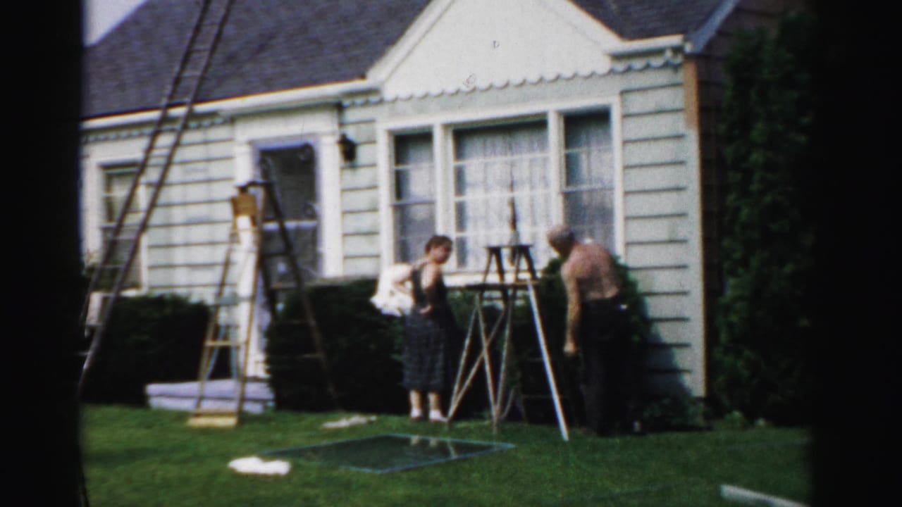 Two men work near ladders outside a light-sided house on a sunny lawn.