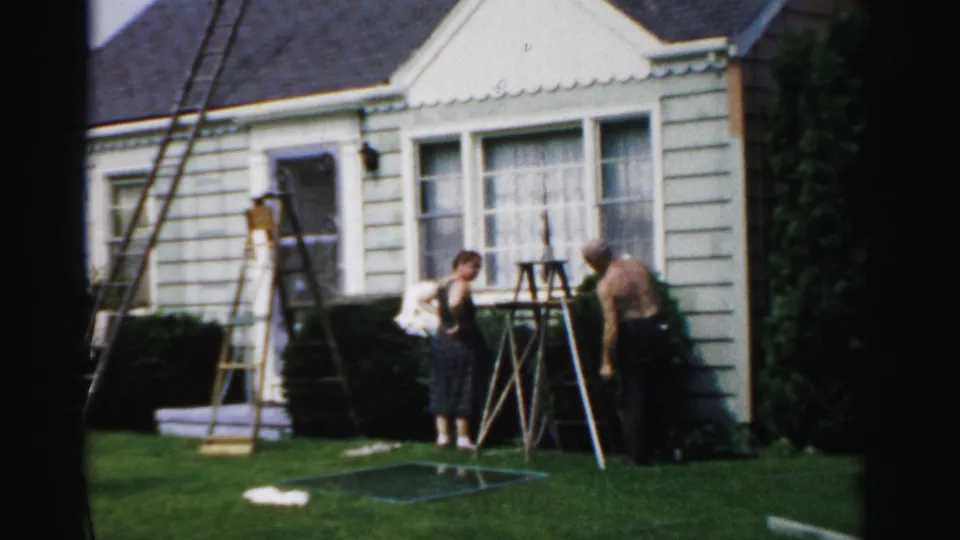In Catskill, New York in 1957, two older men work near ladders outside a small light-sided house on a sunny lawn.