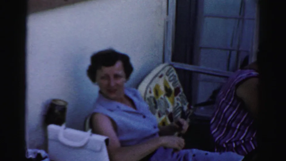 A woman in a sleeveless light top smiles toward the camera while seated outdoors beside another woman near a house wall.