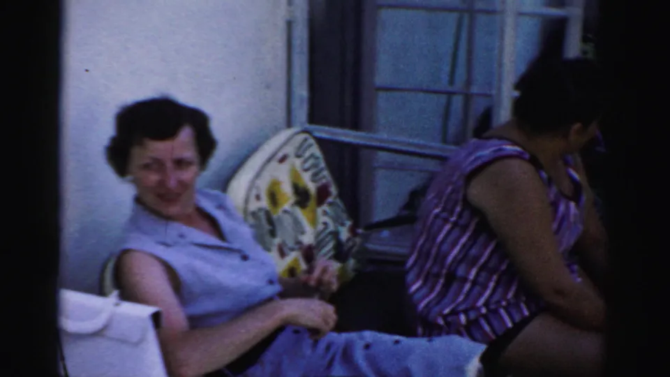 Two women sit outdoors near an open window, with one smiling toward the camera and another in a striped sleeveless top turned to the side.