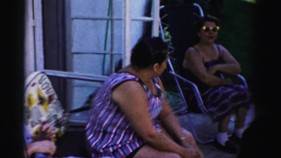 Women sit in outdoor chairs beside the house, with one wearing sunglasses and another turned toward her near ladders or yard equipment.