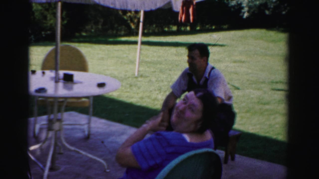 Adults sit on a shaded patio beside a round table and umbrella with a green lawn behind them.
