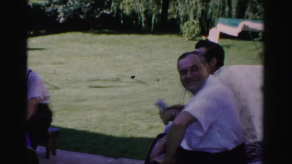 A man in a white shirt smiles toward the camera during an informal 1957 Catskill patio gathering beside a green lawn.