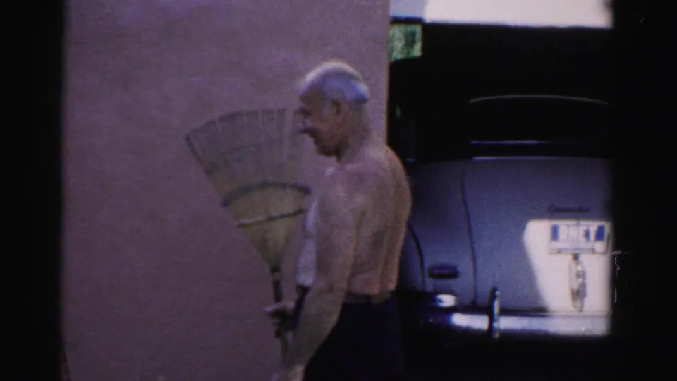 A shirtless older man holds a rake beside a wall and parked vintage car in the 1957 Catskill home-movie scene.