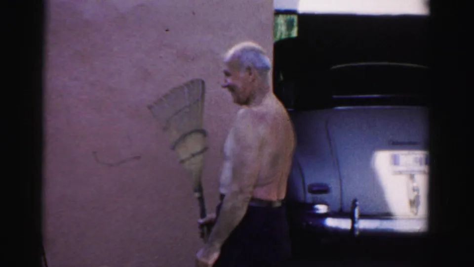 1957 Catskill home-movie frame showing a shirtless man in dark pants holding a rake near a wall and vintage car.