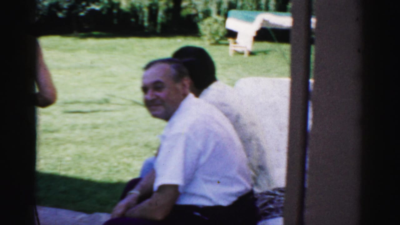 1957 Catskill home-movie frame with a seated man in a white shirt looking toward the camera beside a green lawn.