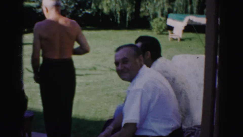 1957 Catskill frame showing a shirtless man from behind as seated men remain near the porch and lawn.