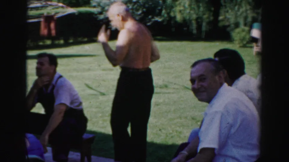 1957 Catskill home-movie frame with a shirtless man holding a rake on the lawn near seated men and trees.