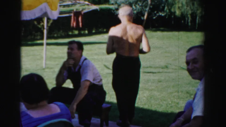 1957 Catskill home-movie frame showing adults gathered on a green backyard lawn while a shirtless older man stands nearby.