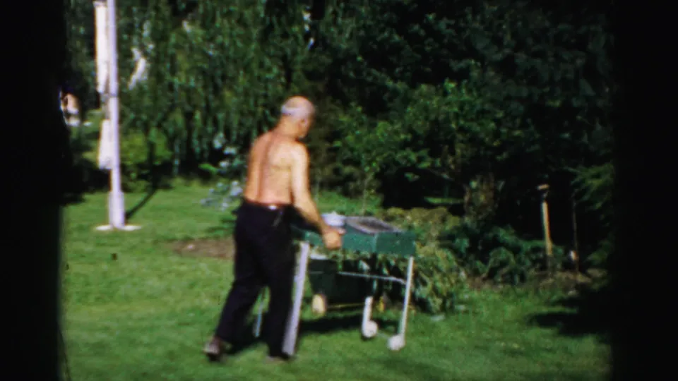 In a Catskill backyard, a shirtless older man approaches a small outdoor table on the grass near dense trees.