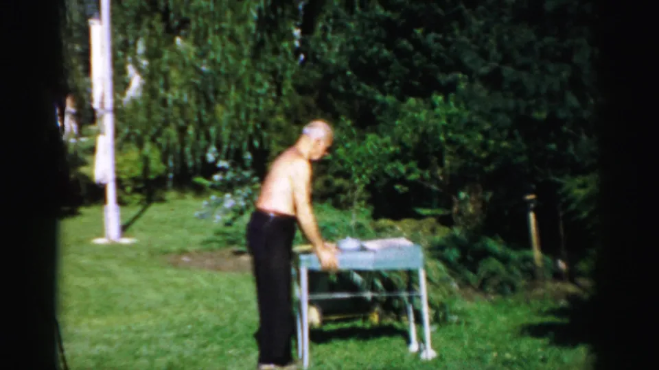 The older man leans over a small table in the 1957 Catskill backyard, with trees and lawn filling the frame.