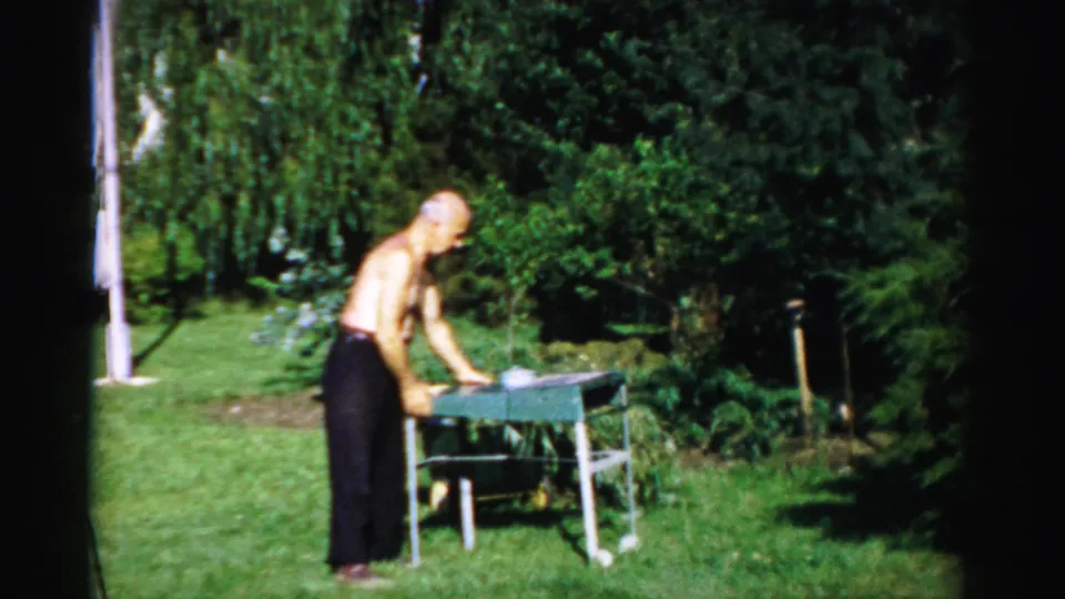 The man continues setting up a small table on the Catskill lawn, framed by trees and strong midday light.