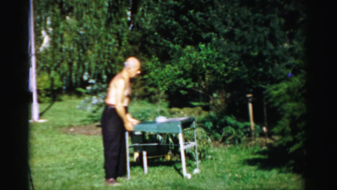 In 1957 Catskill, New York home-movie footage, an older shirtless man adjusts a small green grill on a lawn.