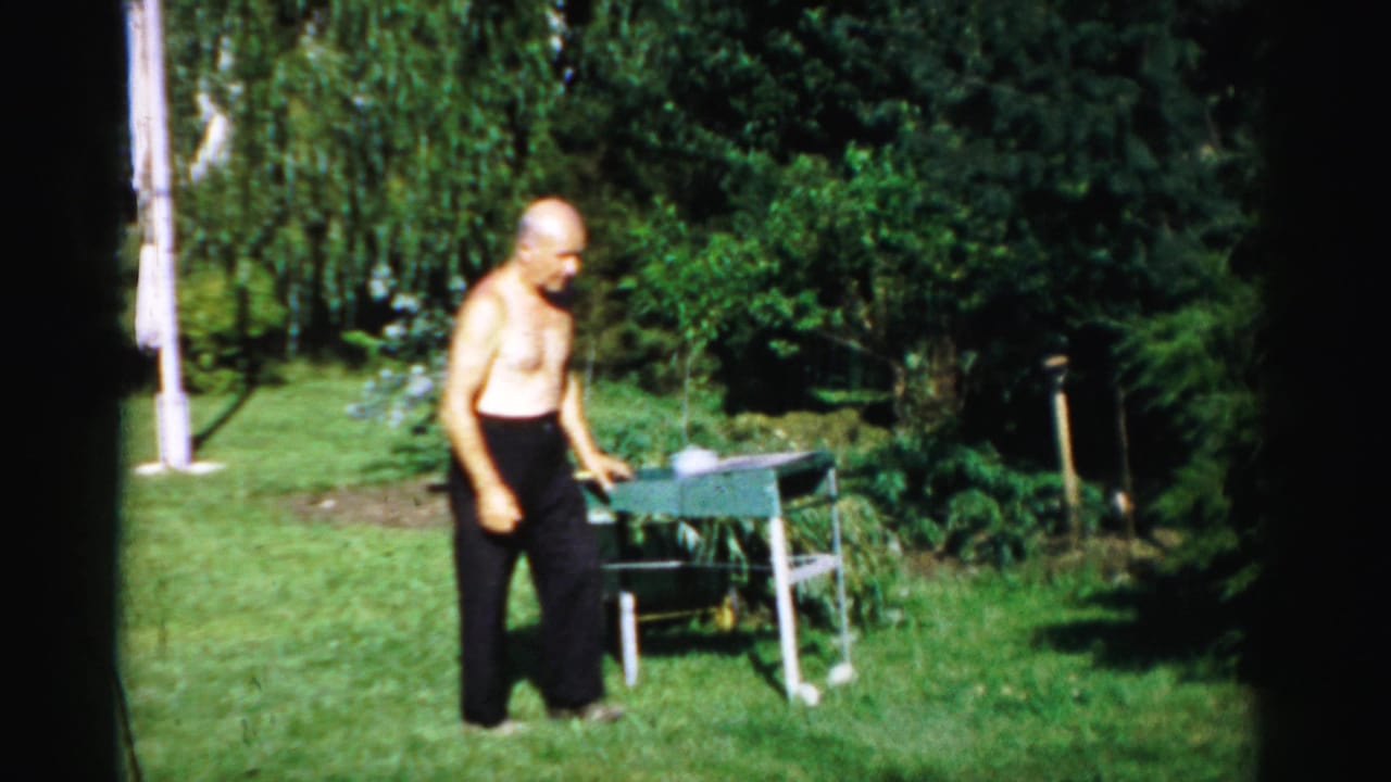 The man stands near the small outdoor grill in a sunlit Catskill backyard, with trees and shrubs behind him.