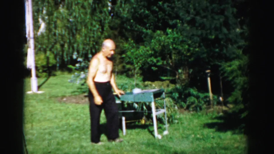 The man stands near the small outdoor grill in a sunlit Catskill backyard, with trees and shrubs behind him.