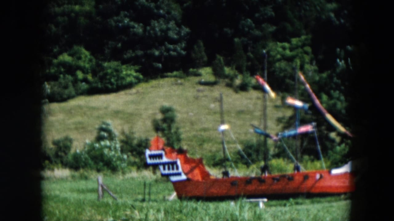 1957 Hicksville home-movie frame showing a red miniature pirate ship displayed in a grassy field with trees in the background.