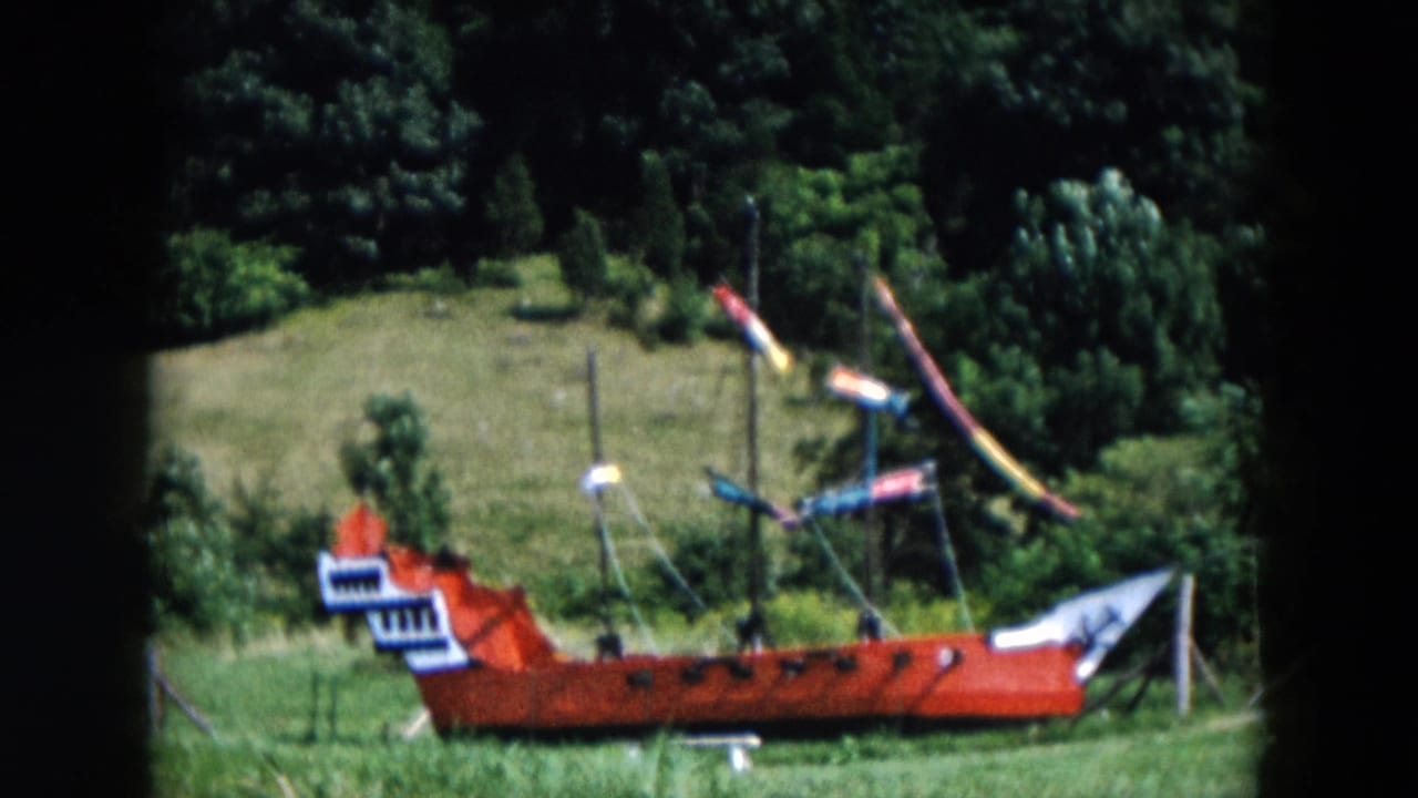 The red pirate ship model appears in profile on grass, with multiple masts, small flags, and dense trees beyond the field.