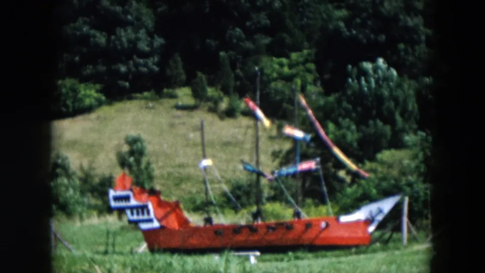 The red pirate ship model appears in profile on grass, with multiple masts, small flags, and dense trees beyond the field.