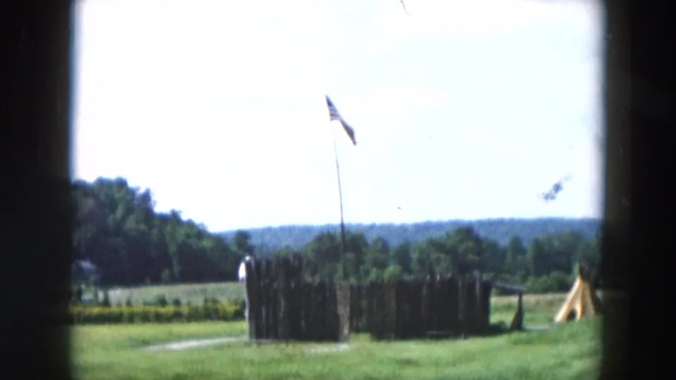 A 1957 Hicksville outdoor frame shows a tall flagpole near a low fenced enclosure, grass, trees, and distant hills.