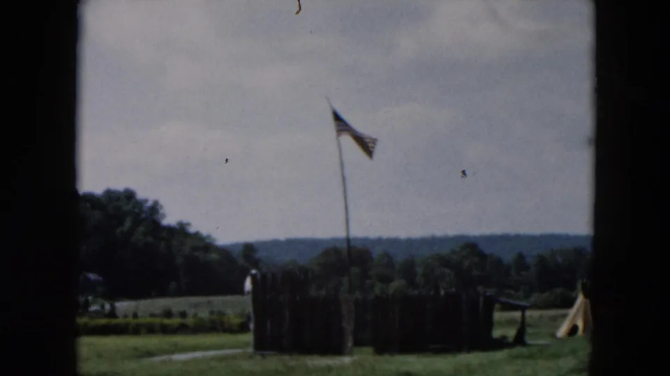 A darker 8mm frame shows the flagpole and fenced display area from a distance, with grass, trees, and low hills behind.