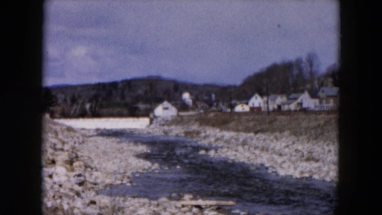 1957 8mm view of a stream flowing through a rocky bed near distant buildings and trees in Hicksville, New York.