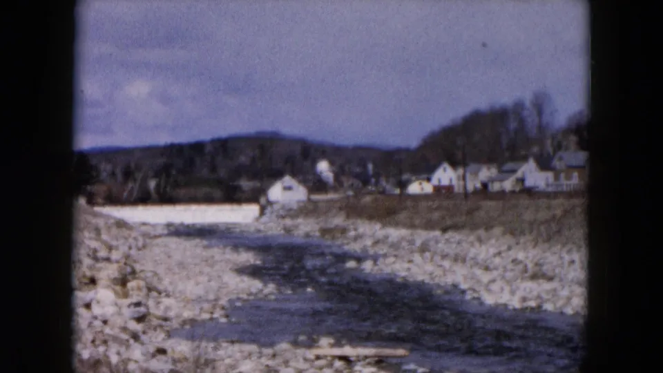 1957 8mm view of a stream flowing through a rocky bed near distant buildings and trees in Hicksville, New York.